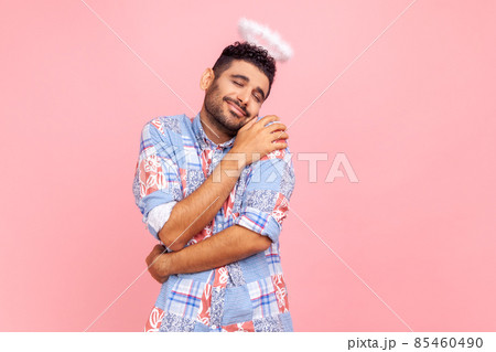 Portrait of amazing angelic man with beard and halo over head smiling contentedly and hugging himself, self-love, support, keeps eyes closed. Indoor studio shot isolated on pink background. Portrait of amazing angelic man with beard and halo over head smiling contentedly and hugging himself, self-love, support, keeps eyes closed. Indoor studio shot isolated on pink background. 85460490