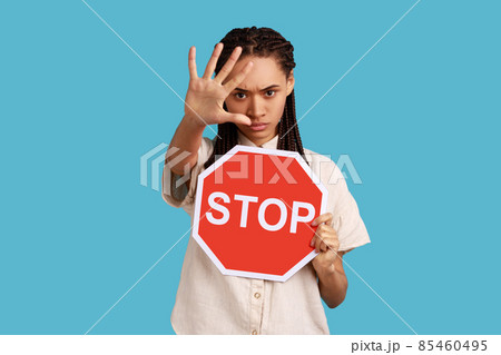 Access denied. Bossy woman with dreadlocks showing stop gesture with hand and holding red stop sign board, enter is restricted, wearing white shirt. Indoor studio shot isolated on blue background. 85460495