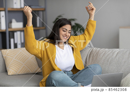 Overjoyed arab lady sitting on sofa with laptop, gesturing YES, celebrating success at home, free space Overjoyed arab lady sitting on sofa with laptop, gesturing YES, celebrating success at home, free space 85462308