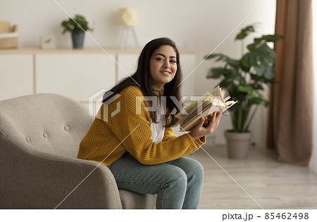 Peaceful stay home pastimes. Happy arab lady reading book, sitting on armchair in living room and smiling at camera Peaceful stay home pastimes. Happy arab lady reading book, sitting on armchair in living room and smiling at camera 85462498