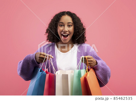 Beautiful millennial black woman looking inside bright shopping bags, happy with her purchases over pink background Beautiful millennial black woman looking inside bright shopping bags, happy with her purchases over pink background 85462535