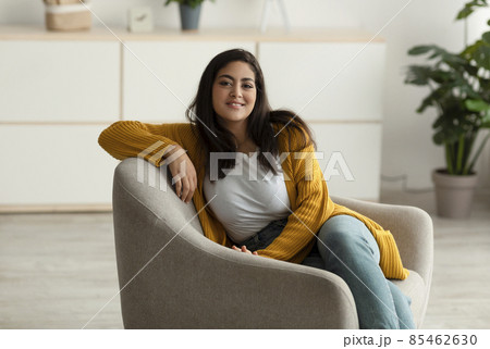 Happy arab lady sitting in comfy armchair and smiling at camera, enjoying lazy morning, having peaceful weekend at home 85462630