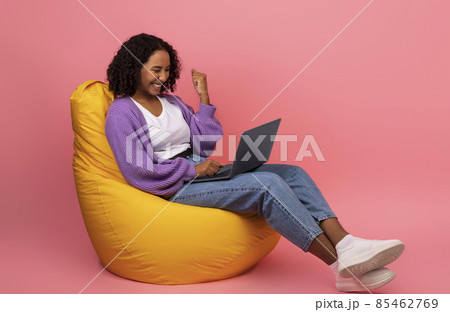 Online win. Excited young black woman sitting in bean bag chair with laptop, making YES gesture, achieving success Online win. Excited young black woman sitting in bean bag chair with laptop, making YES gesture, achieving success 85462769