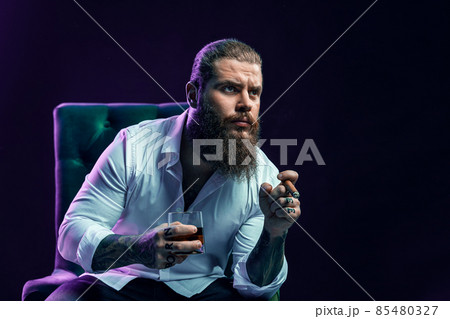 Portrait of thinking bearded man who smoke cigar holds glass of whiskey while sittingin armchair. Portrait of thinking bearded man who smoke cigar holds glass of whiskey while sittingin armchair. 85480327