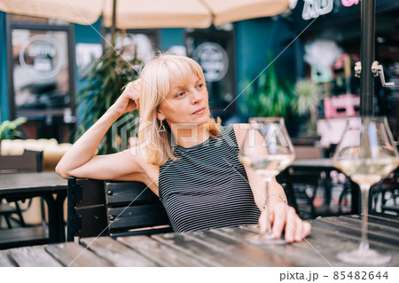 Thoughtful adult mature woman sitting in bar outdoors with wine glasses and blurry restaurant background scene, drinking white wine. Summer sunny day on patio. People lifestyle. 85482644