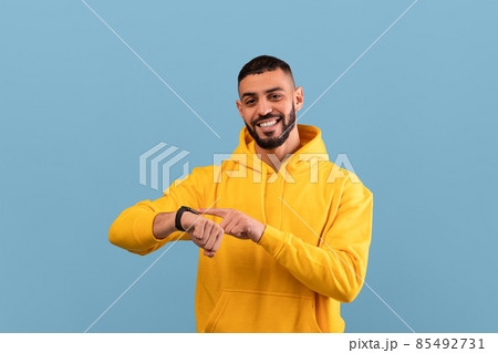 Happy middle eastern guy touching his smartwatch and smiling at camera, standing over blue studio background 85492731