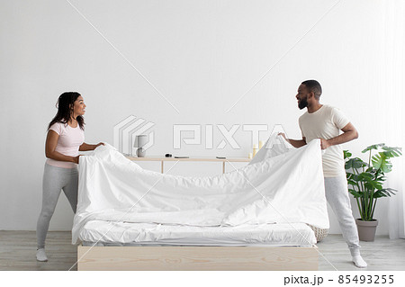 Cheerful young black woman and guy make bed and hold blanket in bedroom interior 85493255