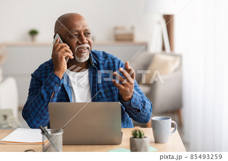 Senior African Businessman Talking On Phone Sitting At Laptop Indoor Senior African Businessman Talking On Phone Sitting At Laptop Indoor 85493259