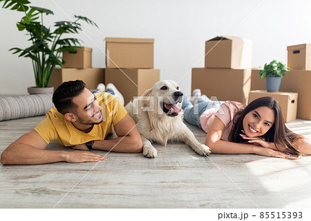 Cool young diverse couple lying on floor among carton boxes with their cute dog, relocating to new home with pet 85515393