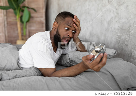 Shocked black man looking at clock in bed Shocked black man looking at clock in bed 85515529
