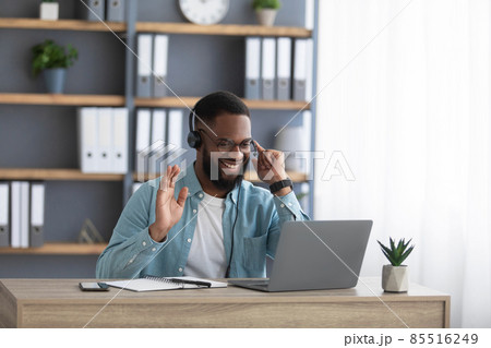Friendly happy young african american businessman with beard in glasses and headphones works on pc and waves his hand Friendly happy young african american businessman with beard in glasses and headphones works on pc and waves his hand 85516249