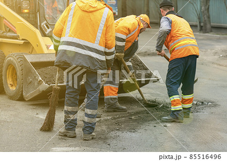 Road workers in bright orange reflective uniforms use shovels to scrape accumulated sand. maintenance of road and highway pavements. pothole repairing works. toned 85516496