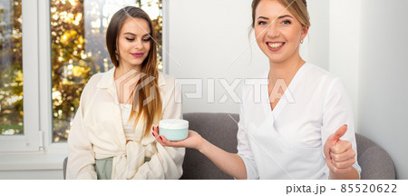Beautician offering product for young woman, shows thumb up, holding a white plastic jar with a cream, mockup, copy space. Beautician offering product for young woman, shows thumb up, holding a white plastic jar with a cream, mockup, copy space. 85520622