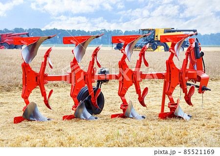 A metal multi-row plow of red color on a blurred background of wheat harvesting with large combines. 85521169
