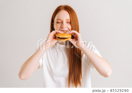 Close-up portrait of pretty young woman with closed eyes enjoying bite of appetizing delicious hamburger on white isolated background in studio. 85529290