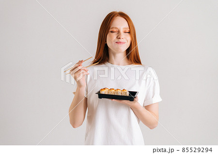 Studio portrait of satisfied young woman with closed eyes eating delicious sushi rolls with chopsticks standing on white isolated background. 85529294