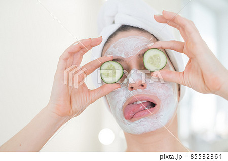 Cheerful woman with a towel on her hair and in a clay face mask fooling around with cucumbers in her hands. Taking care of beauty at home 85532364
