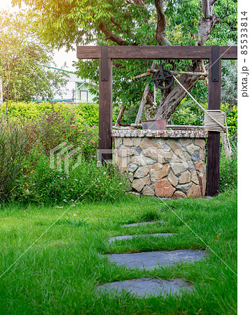 Beautiful artesian well made by stones and wheel pulley with metal bucket and rope in peaceful garden atmosphere. Retro stone water well in rural area. Garden decoration with antique items. 85533814