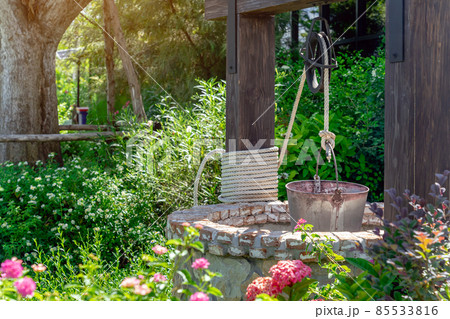 Beautiful artesian well made by stones and wheel pulley with metal bucket and rope in peaceful garden atmosphere. Retro stone water well in rural area. Garden decoration with antique items. 85533816