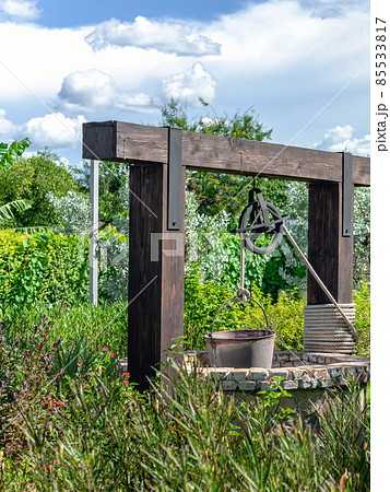 Beautiful artesian well made by stones and wheel pulley with metal bucket and rope in peaceful garden atmosphere. Retro stone water well in rural area. Garden decoration with antique items. 85533817