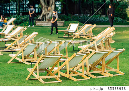 Many empty white deck chairs with tables in lawn is surrounded by shady green grass. Comfortable on outdoor patio chairs in garden.  Lawn chairs in the park. Sunbeds in the garden. Selective focus. 85533958