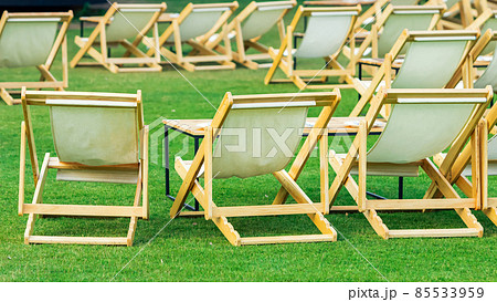 Many empty white deck chairs with tables in lawn is surrounded by shady green grass. Comfortable on outdoor patio chairs in garden.  Lawn chairs in the park. Sunbeds in the garden. Selective focus. 85533959