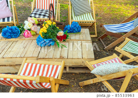Beautiful artificial bouquets of various colors placed on wooden table among deck chairs in garden. Multi-colored artificial flowers adorn the wooden table for beautiful and relaxing. Selective focus. 85533963