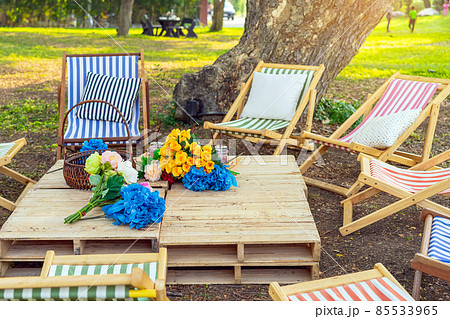 Beautiful artificial bouquets of various colors placed on wooden table among deck chairs in garden. Multi-colored artificial flowers adorn the wooden table for beautiful and relaxing. Selective focus. 85533965