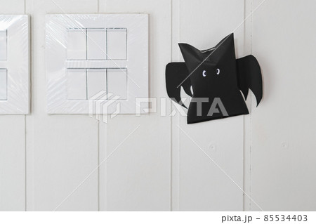 Origami bat made of black paper near light switches on white wall background. Dark paper ghost Halloween party concept origami paper bat. The figure of black paper bat flying over white background. Origami bat made of black paper near light switches on white wall background. Dark paper ghost Halloween party concept origami paper bat. The figure of black paper bat flying over white background. 85534403