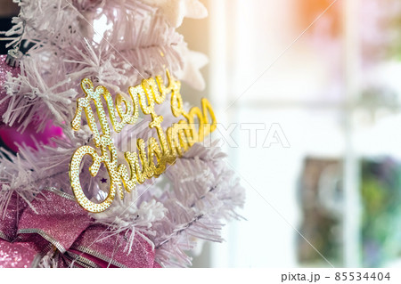 Golden shiny Merry Christmas handwriting logo on white paper tree for christmas time in coffee shop. Christmas tree decorations with color lights and a merry christmas logo in cafe. Selective focus Golden shiny Merry Christmas handwriting logo on white paper tree for christmas time in coffee shop. Christmas tree decorations with color lights and a merry christmas logo in cafe. Selective focus 85534404
