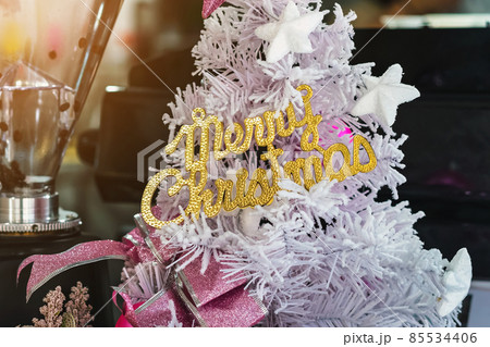 Golden shiny Merry Christmas handwriting logo on white paper tree for christmas time in coffee shop. Christmas tree decorations with color lights and a merry christmas logo in cafe. Selective focus Golden shiny Merry Christmas handwriting logo on white paper tree for christmas time in coffee shop. Christmas tree decorations with color lights and a merry christmas logo in cafe. Selective focus 85534406