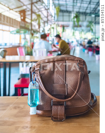 Mini portable blue alcohol gel bottle to kill Corona Virus(Covid-19) hang on brown leather shoulder bag of a woman on table at cafeteria. New normal lifestyle. Selective focus on alcohol gel. Mini portable blue alcohol gel bottle to kill Corona Virus(Covid-19) hang on brown leather shoulder bag of a woman on table at cafeteria. New normal lifestyle. Selective focus on alcohol gel. 85534511