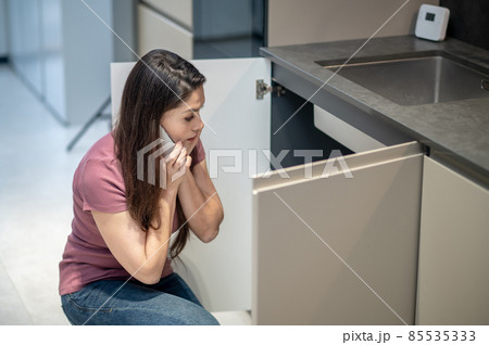 Confused woman with smartphone near ear looking under sink Confused woman with smartphone near ear looking under sink 85535333