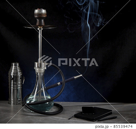 Beautiful modern hookah, from the mouthpiece of which smoke rises, stands on a table surrounded by a metal protective cap and a diary, on a dark background. Copy space. Beautiful modern hookah, from the mouthpiece of which smoke rises, stands on a table surrounded by a metal protective cap and a diary, on a dark background. Copy space. 85539474