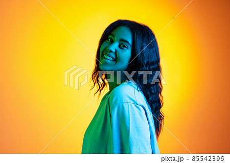 Happy young pretty dark skinned girl wearing white t-shirt isolated on yellow background in neon light, filter. Happy young pretty dark skinned girl wearing white t-shirt isolated on yellow background in neon light, filter. 85542396