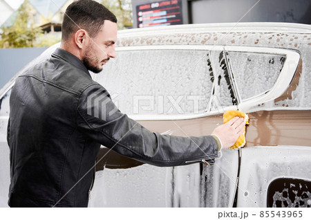 Back view of young man washing car on carwash station outdoor. Handsome worker cleaning automobile, using sponge and soap. 85543965