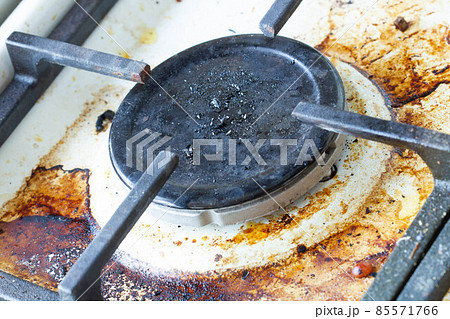 Dirty gas stove surface. Gas burner and cast iron grate of a gas oven surrounded by old leftovers of food and drinks. Top area surface and burner heads needs cleaning from burnt-on grease. Close-up Dirty gas stove surface. Gas burner and cast iron grate of a gas oven surrounded by old leftovers of food and drinks. Top area surface and burner heads needs cleaning from burnt-on grease. Close-up 85571766