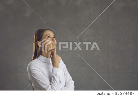 Portrait of a dreamy and pensive woman standing thoughtfully on a gray background. Young beautiful woman looks up and touches her chin, pondering a proposal or making a choice. Place for text. 85574487