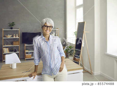 Portrait of a beautiful, stylish and cheerful senior woman looking at the camera. Woman with gray hair and glasses is posing relaxed at home or in the office with a natural face and a confident smile. Portrait of a beautiful, stylish and cheerful senior woman looking at the camera. Woman with gray hair and glasses is posing relaxed at home or in the office with a natural face and a confident smile. 85574488