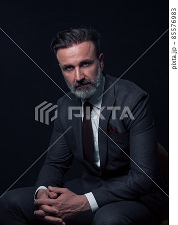 Portrait of adult businessman wearing trendy suit and sitting in modern studio on stylish chair against the black background Portrait of adult businessman wearing trendy suit and sitting in modern studio on stylish chair against the black background 85576983