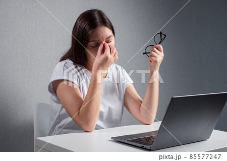 young woman massaging the bridge of nose, holding glasses in other hand, sitting in front of a laptop young woman massaging the bridge of nose, holding glasses in other hand, sitting in front of a laptop 85577247