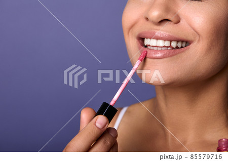 Close-up. Cropped view of attractive smiling woman with perfect clean healthy skin applying lip gloss hygienic balm on plump lips isolated on purple background. Copy ad space. Make-up concept 85579716