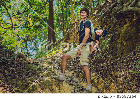 The father carries his son in a baby carrying is hiking in the forest. Tourist is carrying a child on his back in the nature of Vietnam 85597264