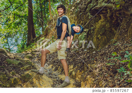 The father carries his son in a baby carrying is hiking in the forest. Tourist is carrying a child on his back in the nature of Vietnam The father carries his son in a baby carrying is hiking in the forest. Tourist is carrying a child on his back in the nature of Vietnam 85597267