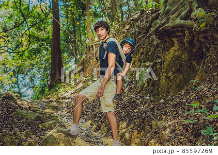 The father carries his son in a baby carrying is hiking in the forest. Tourist is carrying a child on his back in the nature of Vietnam The father carries his son in a baby carrying is hiking in the forest. Tourist is carrying a child on his back in the nature of Vietnam 85597269