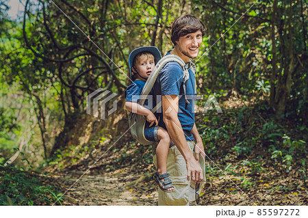 The father carries his son in a baby carrying is hiking in the forest. Tourist is carrying a child on his back in the nature of Vietnam The father carries his son in a baby carrying is hiking in the forest. Tourist is carrying a child on his back in the nature of Vietnam 85597272