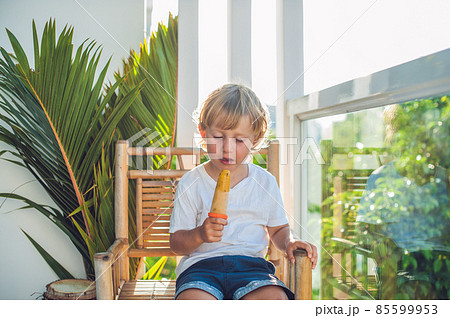 Cute little blond boy eating a homemade icecream sitting on a wooden chair. 85599953