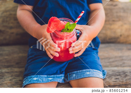 The boy is holding Healthy watermelon smoothie in Mason jar with mint and striped straws on a wood background 85601254