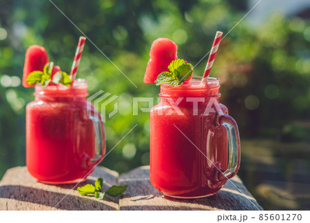 Healthy watermelon smoothie in Mason jars with mint and striped straws against the background of greenery Healthy watermelon smoothie in Mason jars with mint and striped straws against the background of greenery 85601270