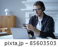 Pensive male entrepreneur in headphones sitting at desk at home office and using mobile phone 85603503